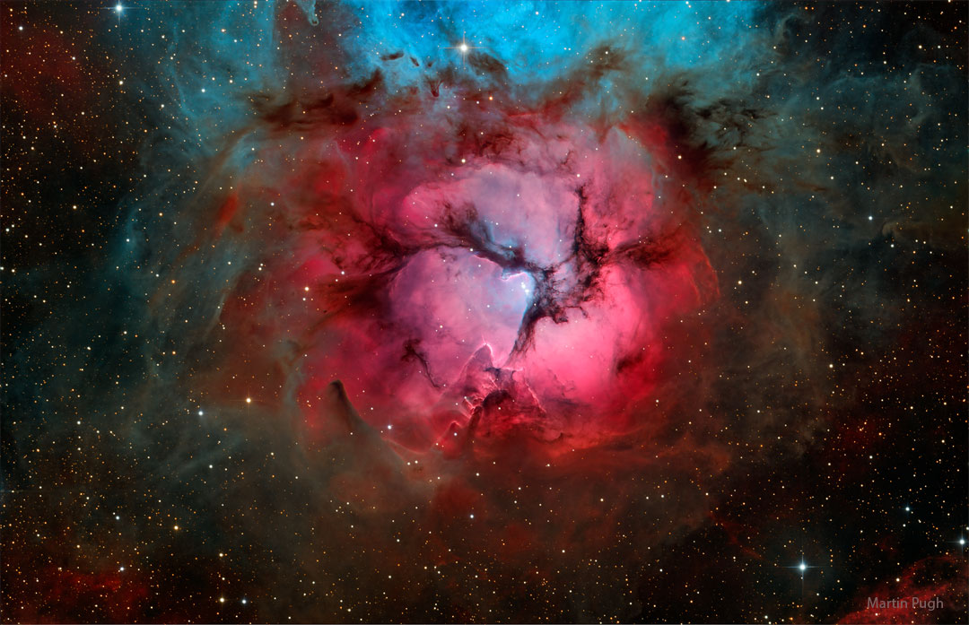 A bright red gaseous nebula is pictures with three dark
dust lanes meeting in the centre. The top of the nebula appears
blue.
Please see the explanation for more detailed information.