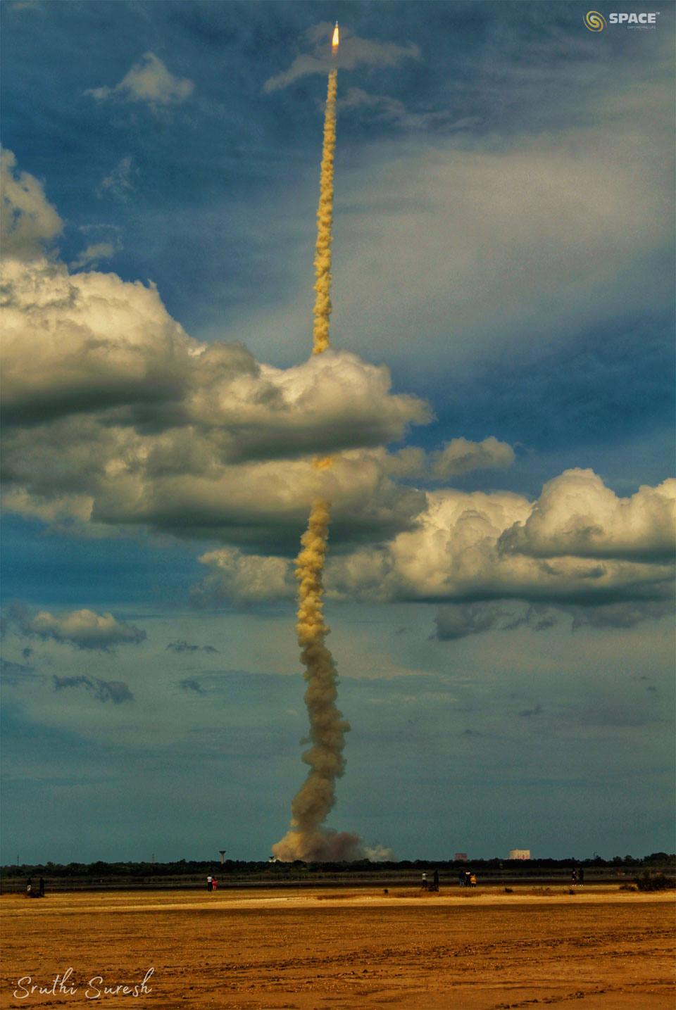 A rocket is seen after lift-off with a long smoke plume.
The rocket is captured against a blue sky and has gone through
a cloud deck. In the foreground is an empty tan-coloured field. 
Please see the explanation for more detailed information.
