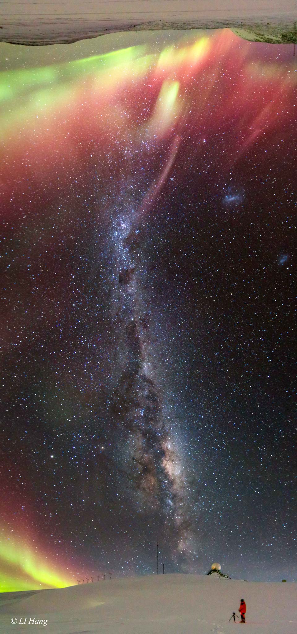 A long vertical image shows a band of the night sky from
horizon at the bottom to the opposite horizon -- at the image top.
A person stands on a snow covered landscape with the central
band of the Milky Way running between horizons. Each horizon
is lit by red, yellow, and green aurorae.
Please see the explanation for more detailed information.