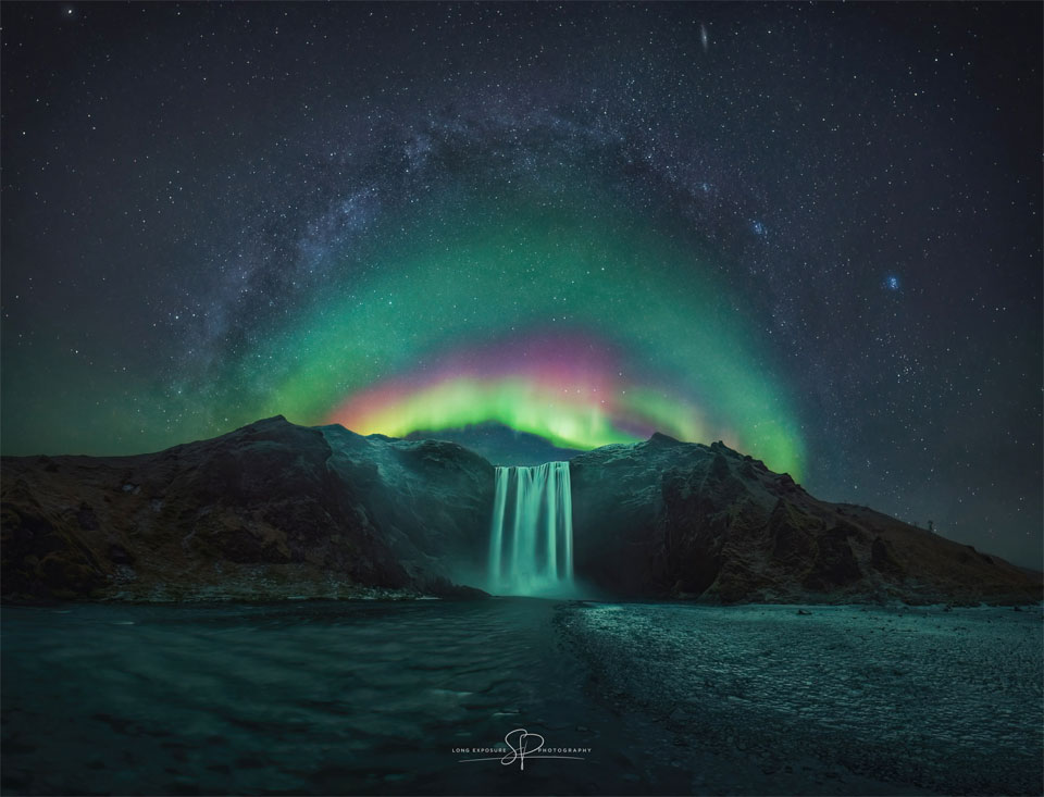 A waterfall is shown in the image centre below a starry
sky. Arching above the waterfall is a colourful aurora. Arching
above the aurora is the central band of the Milky Way. 
Please see the explanation for more detailed information.