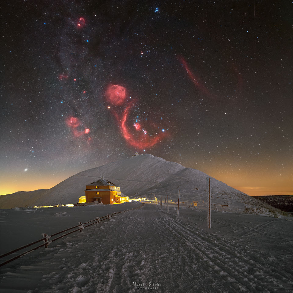 A snowy landscape is pictured with a big hill in the centre.
Above the hill is a starfield with the stars and nebulae of the 
constellation Orion appearing, with the red glow of the 
nebulae in great contrast to the dark sky and bright snow.
Please see the explanation for more detailed information.
