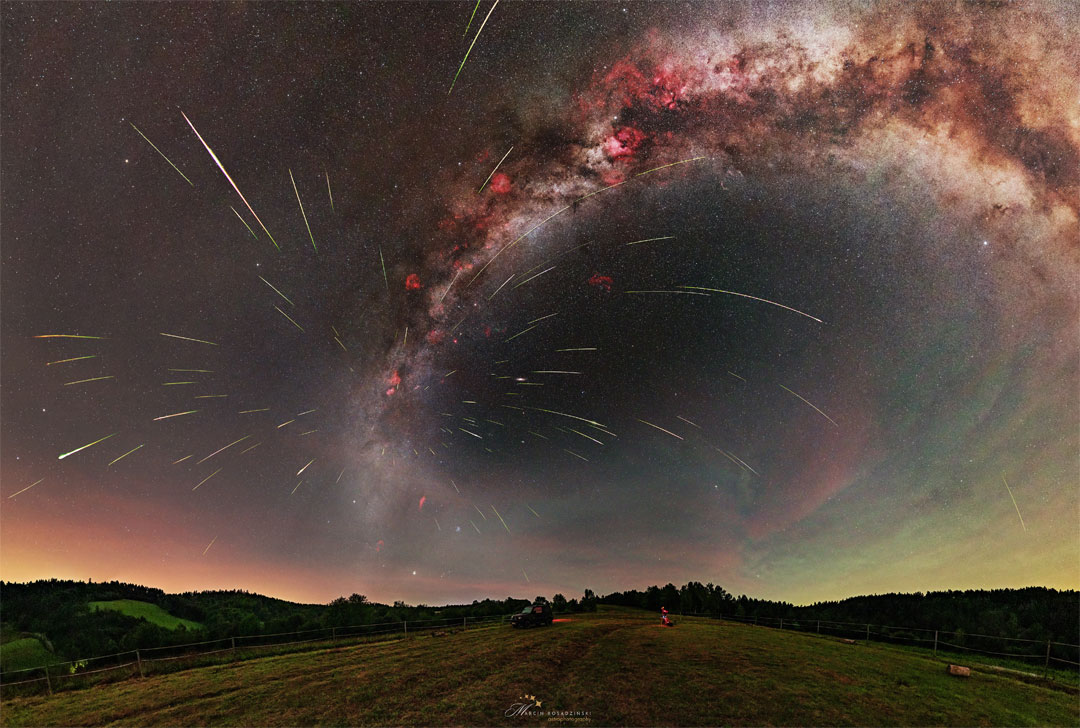 A starfield is shown above a grassy field with hills on
the horizon. The band of our Milky Way Galaxy arches across
toward the right. Many streaks appear emanating out from a 
place on the Milky Way just above the horizon.  
Please see the explanation for more detailed information.