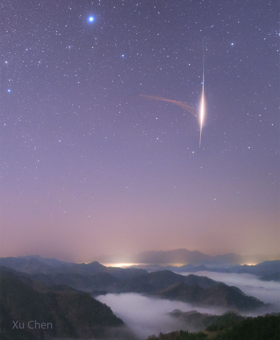 A view of mountains over clouds shows a starfield
with a purple glow. Prominent on the right is the trail
of a bright meteor. To the left of the meteor and 
connecting to the meteor is something unusual: a light
brown triangular puff. 
Please see the explanation for more detailed information.