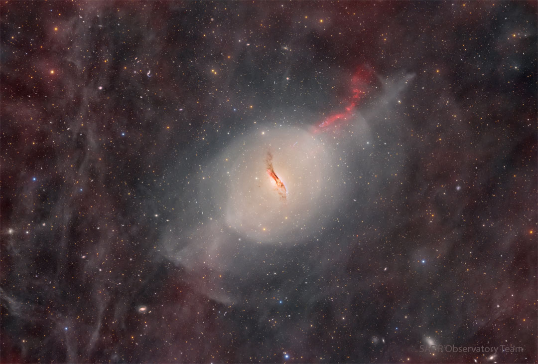 A starfield glows faintly with red glowing gas and dust.
In the centre there is some sort of unusual galaxy with a red jet
emanating toward the upper right and a dark dust lane through
its centre.  
Please see the explanation for more detailed information.