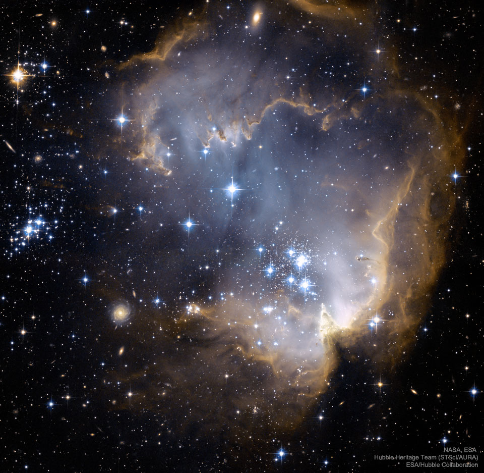 A starfield surrounds a large light-brown nebula that
has several dust pillars. In the centre of the nebula are many
bright blue stars. Some distant galaxies are visible 
through and around the nebula.
Please see the explanation for more detailed information.