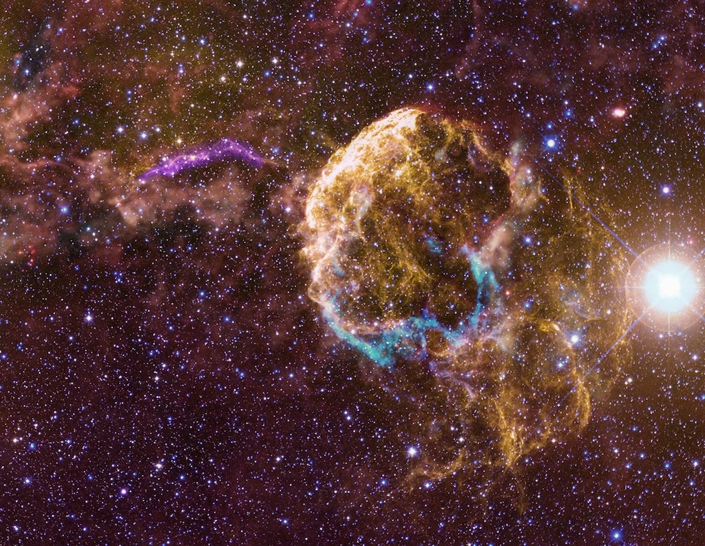 Against a bright field of stars, a yellow nebula with blue details,
	  a purple filament on the left and a bright star on the right.