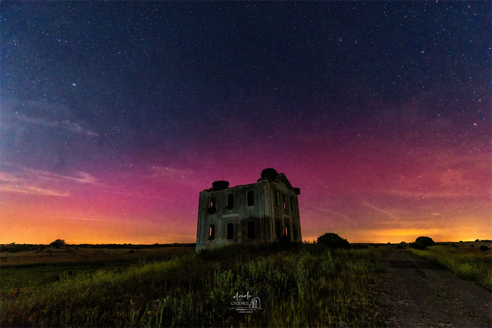 A dark rectangular building is seen across a green field
with colourful aurora, mostly red, seen in the background sky. 
Past the aurora, the sky is also filled with stars. 
Please see the explanation for more detailed information.