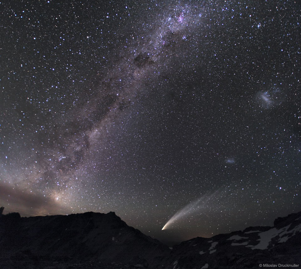 A rocky landscape is capped by a dark night sky. In the 
sky, the band of our Milky Way Galaxy runs along the right,
while two fuzzy patches that are the LMC and SMC are visible on
the right. Thousands of stars are resolved all over the frame. 
Please see the explanation for more detailed information.