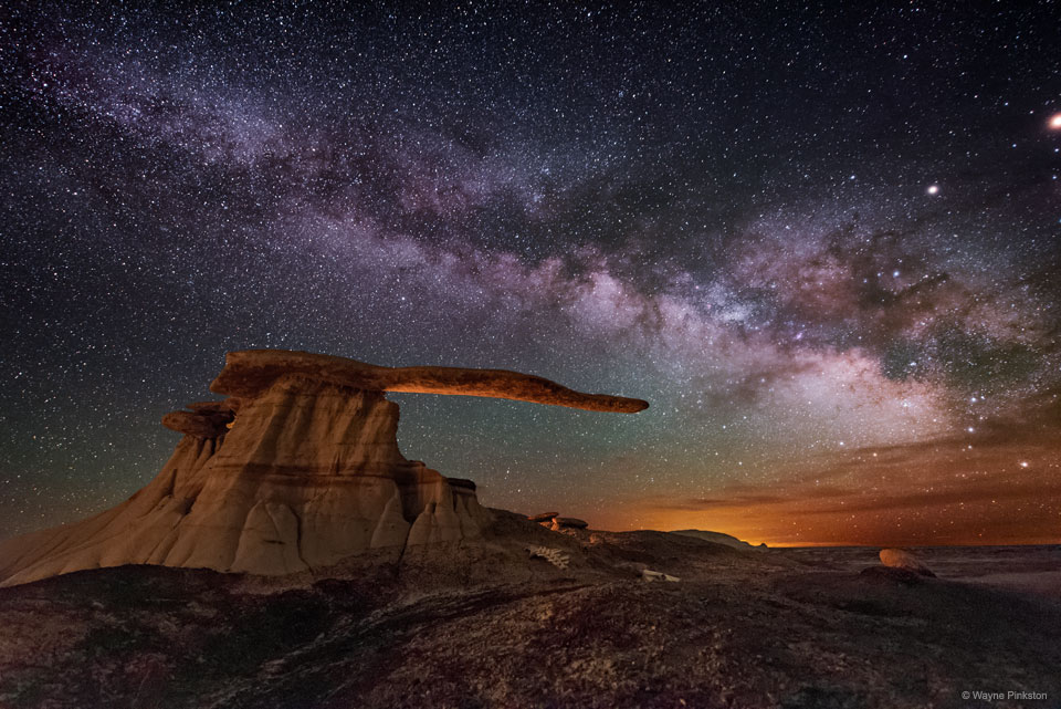 The band of the Milky Way runs across a night sky filled 
with stars. Colourful clouds are on the right horizon. A strange
rock structure appears in the image centre with a base and 
an extended arm that seems to point to the colourful horizon.
Please see the explanation for more detailed information.