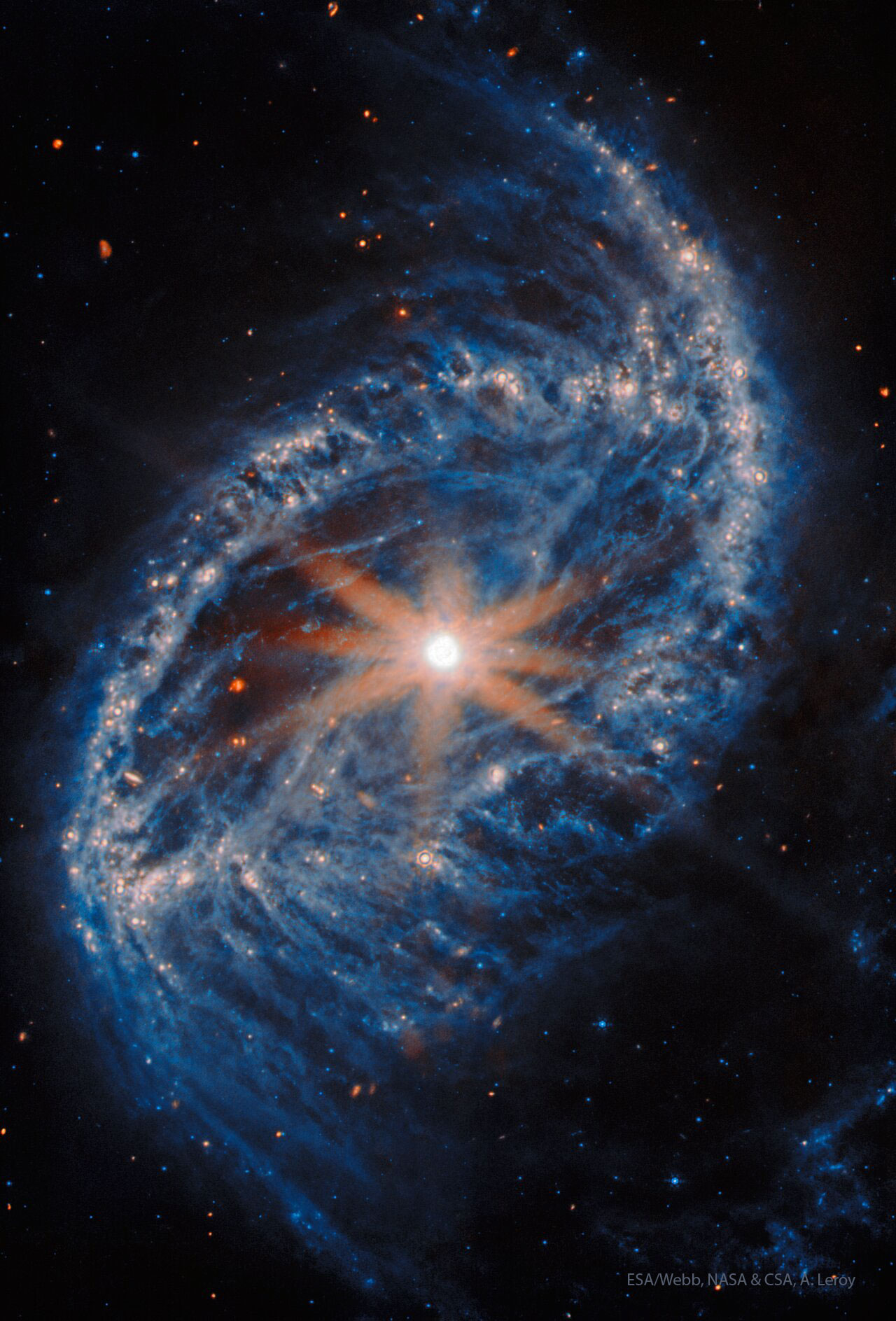 An oval galaxy is shown against a field of stars.
The outer rings shows many bright blue stars. In the 
centre is a bright nucleus with eight spikes jutting out.
Please see the explanation for more detailed information.