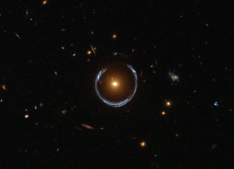 A dark star field with several small galaxies 
features a yellowish galaxy in the centre. Surrounding
this galaxy is a blue nearly circular horseshoe shaped
object. 
Please see the explanation for more detailed information.