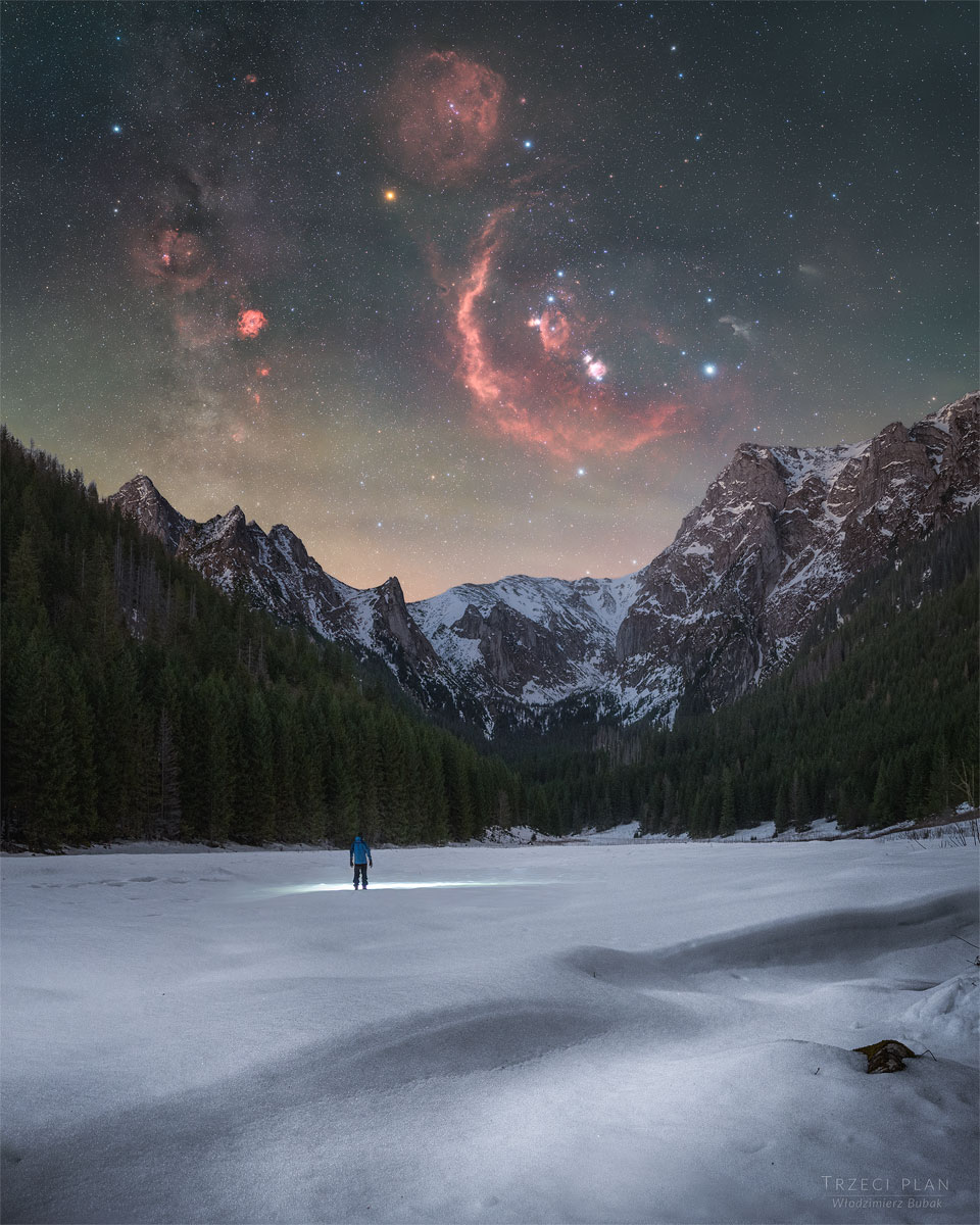 A person stands in the distance on a snowy landscape shining
a flashlight on the ground. Trees and snow capped mountains are
on the horizon. In the starry sky above, several iconic red
nebulae are visible. 
Please see the explanation for more detailed information.