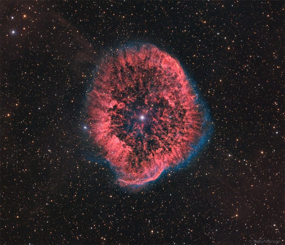 A red oval and textured nebula is seen surrounded by a faint
blue glow. A bright star is visible in the centre, and many faint
stars are visible in the background. 
Please see the explanation for more detailed information.