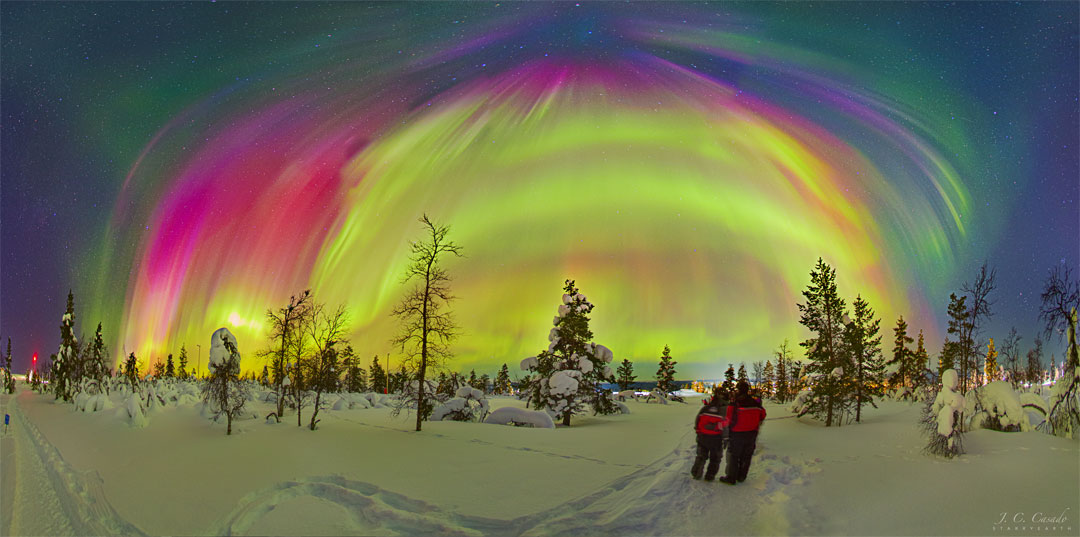 Two people dressed in red coats are standing on a snowy
landscape with bare trees. Above, many aurorae of different 
colours appear, with some stars visible in the background.
Please see the explanation for more detailed information.