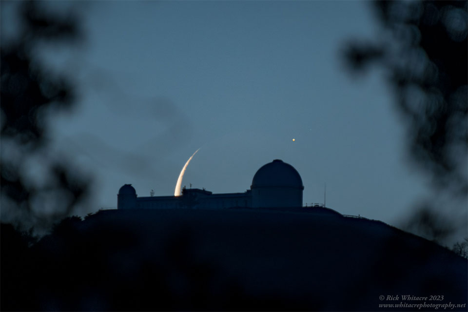 A dark mountain lies in the centre with an observatory
building sporting two telescope domes. The background sky
appears dark blue. Behind the centre of the observatory
is part of a crescent moon, with an unusual bright spot
to its upper left.
Please see the explanation for more detailed information.