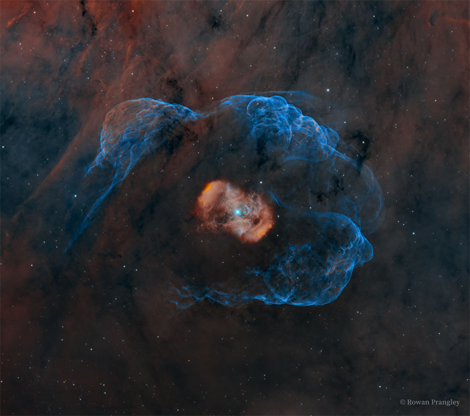 A blue star is seen in the centre of a red nebula 
itself surrounded by a faint blue nebula. The surrounding
starfield itself has a faint red-brown emission clouds.
Please see the explanation for more detailed information.