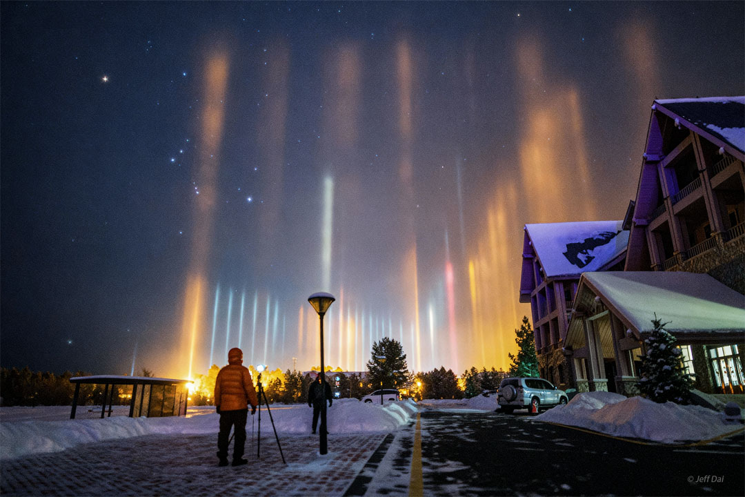 A starry sky appears above a snowy street with a 
house on the right. The constellation of Orion is 
visible on the left. Up from the ground many pillars
of light are seen. Two people stand watching the pillars
on the street in the foreground. 
Please see the explanation for more detailed information.