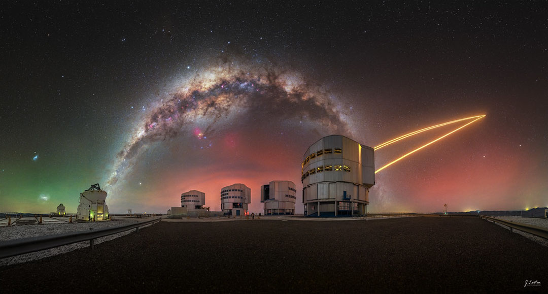 A colourful sky appears above several large telescopes.
Prominent in the sky is the band of the Milky Way Galaxy which
arches across the frame. Orange laser beams connect one
of the telescopes to the sky on the image right. 
Please see the explanation for more detailed information.