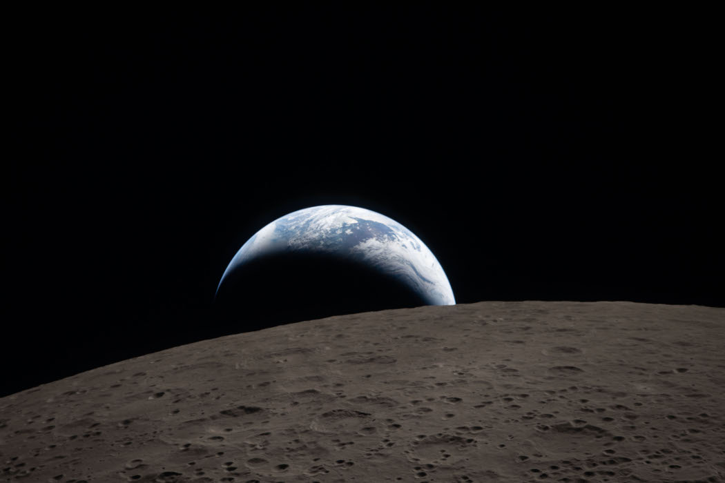 NASA alt text: Earthset captured through the Orion spacecraft window at 6:41 p.m. EDT, April 6, 2026, during the Artemis II crew’s flyby of the Moon. A muted blue Earth with bright white clouds sets behind the cratered lunar surface. The dark portion of Earth is experiencing nighttime. On Earth’s day side, swirling clouds are visible over the Australia and Oceania region. In the foreground, Ohm crater has terraced edges and a flat floor interrupted by central peaks. Central peaks form in complex craters when the lunar surface, liquefied on impact, splashes upwards during the crater’s formation.