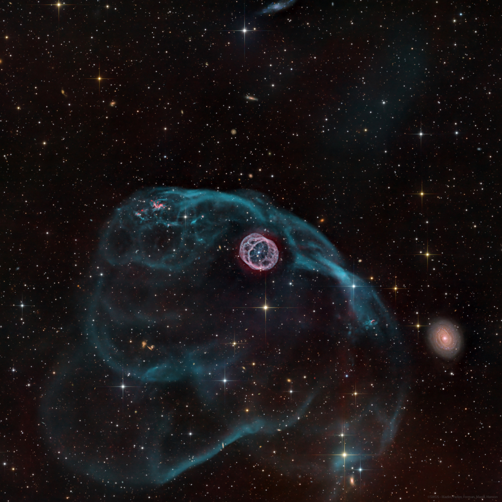 A gaseous structure that resembles a hamster wheel sits near the centre of the image. Inside is a bright white dwarf star. There is a larger asymmetric gas bubble surrounding the inner nebula. A galaxy sits to the bottom right. The background is composed of foreground and background stars, as well as distant galaxies.