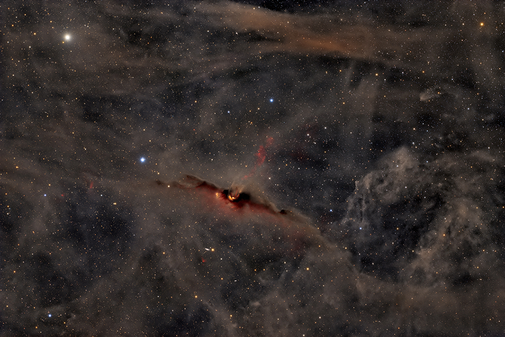 There is a dark cloud of gas and dust resembling a bat with outstretched wings in the middle of the image. Two spots appear to glow from within where there are young stars. A different more diffuse glow appears
to outline just below the bat and extending outward from the bats head. The background is scattered with stars.