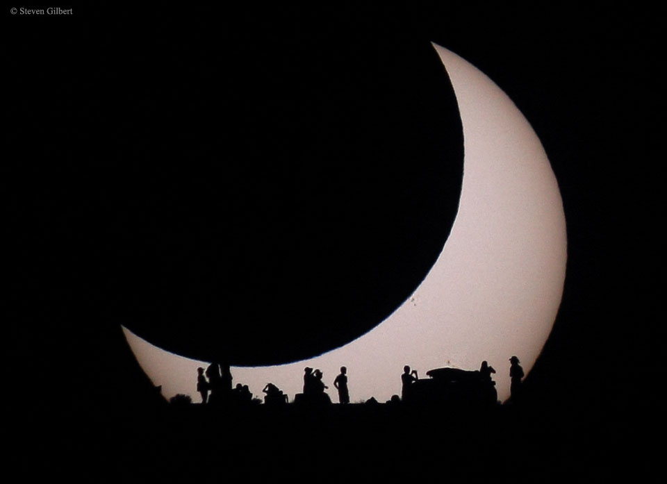A light disk is visible on the lower left but the
rest of the disk is eclipsed by a darker disk of the 
same apparent size. At the bottom of the image are the
silhouettes of several people. 
Please see the explanation for more detailed information.